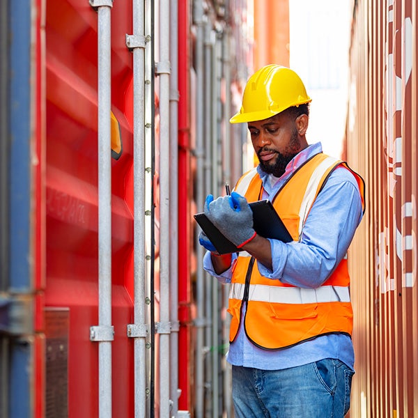 A man in a hard hat and an orange vest is writing notes on his tablet