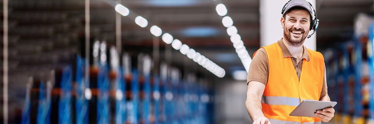 A smiling man in a warehouse who is wearing a headset and an orange vest