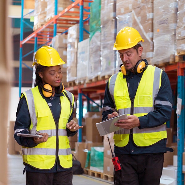 Two coworkers in a storage unit wearing hardhats and yellow vests talking about inventory